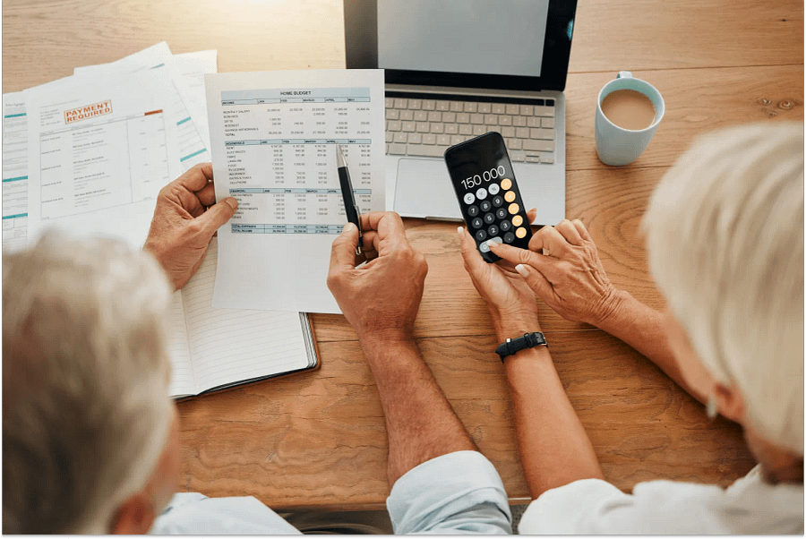 A senior couple calculating their expenses, bills or income during retirement with a calculator, laptop and paper documents from behind.